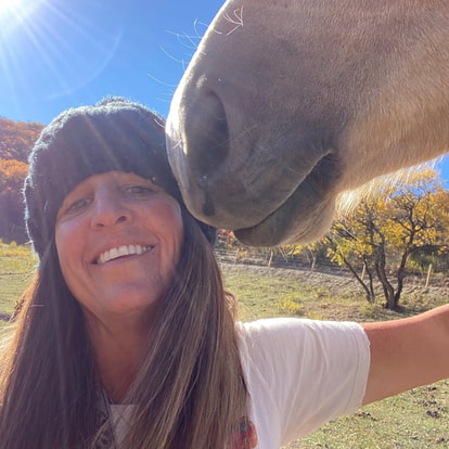 A joyful, and beautiful woman with a grey warm beanie style hat on, smiling at the camera with the muzzle of a previously wild palomino horse against the camera facing left side of her face. her arm is lifted up to the horses neck. She is wearing a white t shirt as well, and it is a sunny fall day- indicated by the color of the leaves on the tree in the background which is yellow. 